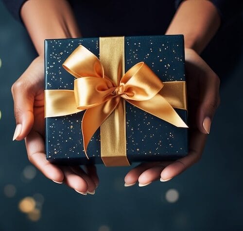 Woman hands holding elegant present gift box with golden ribbon over blue background with confetti. Christmas, new year, valentine’s day, mother day, father day greeting card. Top view